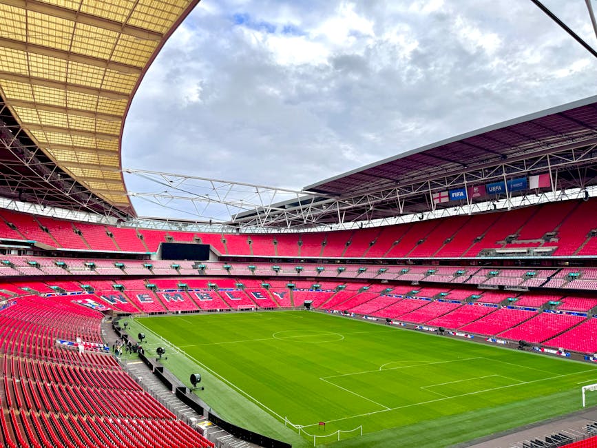 Wide-angle view of Twickenham Stadium's interior, showcasing the vibrant red seating sections arranged in a semi-circular pattern around the green grass pitch. The field is neatly maintained with visible white lines marking the boundaries and goal areas. Overhead, the cloudy sky is partially visible through the open roof structure, which features yellow and purple roofing panels supported by metal trusses. Surrounding the pitch, there are various black lighting and camera equipment positioned on the sidelines. The seating area appears clean and well-organized, with no visible debris or dust, emphasizing the stadium's readiness for an event. The lighting in the image highlights the pristine condition of the surfaces, and the overall scene conveys a sense of modern, well-maintained sports infrastructure, consistent with a professional cleaning and maintenance standard promoted by Twickenham Carpet Cleaning for large venue carpet and surface cleaning services.