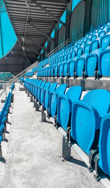 A row of bright blue stadium seats arranged on a concrete tiered floor, with the seats facing towards the playing field of Twickenham Stadium. The seats are made of durable plastic, neatly aligned, and appear clean and well-maintained. The stadium's metal and steel roof structure, painted in dark grey, extends overhead, providing partial shade, and is supported by sturdy beams. Natural daylight illuminates the seating area, highlighting the vibrant colour of the seats and the overall tidy appearance. Alongside the rows, a narrow concrete walkway runs parallel, free of debris or dirt, contributing to the clean and organized environment. This setting exemplifies professional stadium seating, maintained for comfort and hygiene, as part of Twickenham Carpet Cleaning’s surface and deep cleaning services for large-scale events.
