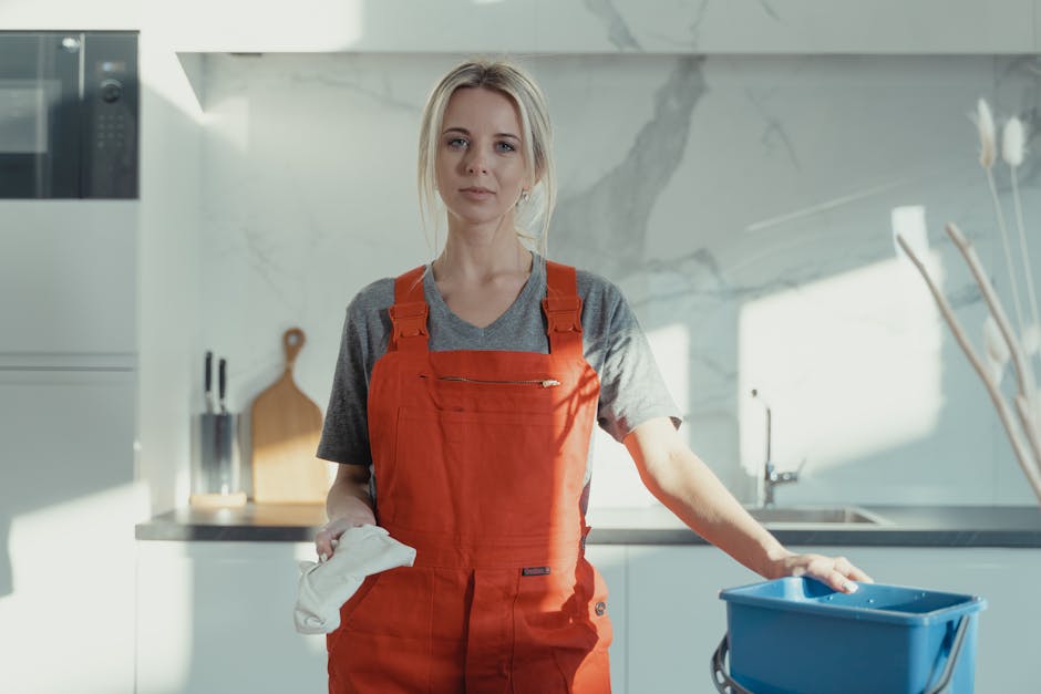 A female cleaning professional wearing a red apron stands in a modern kitchen, holding a white cloth in her left hand and resting her right hand on a blue cleaning bucket. The kitchen features a marble backsplash, stainless steel appliances, and white cabinetry, with natural light illuminating the scene. The surface appears clean and well-maintained, indicating a thorough cleaning process by Twickenham Carpet Cleaning, specializing in domestic surface cleaning and sanitisation for rapid same-day service in Twickenham TW1.