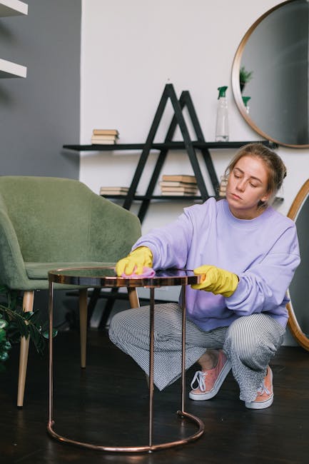 A young woman wearing a lavender sweatshirt, gray checkered pants, yellow cleaning gloves, and white sneakers is kneeling on the dark wooden floor of a modern living room, carefully wiping a round, glass-topped coffee table with a pink cloth. The room features a light-colored wall decorated with a sleek black shelf holding books and small green plants, a large oval mirror, and a small potted plant in the background. A green upholstered armchair is positioned nearby. The space is well-lit with natural light, emphasizing the clean and tidy appearance of the surfaces. The scene showcases domestic cleaning and surface sanitisation, with Twickenham Carpet Cleaning providing comprehensive deep cleaning services.