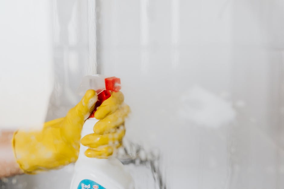 A female cleaning professional wearing a red apron stands in a modern kitchen, holding a white cloth in her left hand and resting her right hand on a blue cleaning bucket. The kitchen features a marble backsplash, stainless steel appliances, and white cabinetry, with natural light illuminating the scene. The surface appears clean and well-maintained, indicating a thorough cleaning process by Twickenham Carpet Cleaning, specializing in domestic surface cleaning and sanitisation for rapid same-day service in Twickenham TW1.