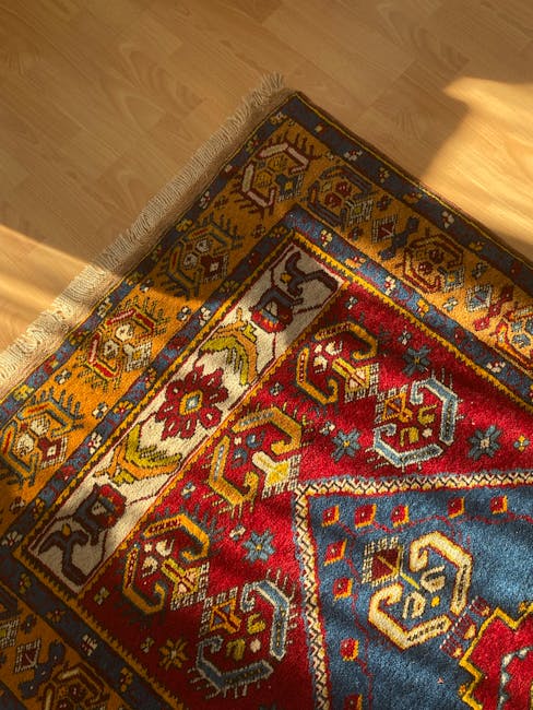 A close-up view of a traditional patterned rug with intricate geometric designs in red, yellow, blue, and white, partially rolled back to reveal the wooden floor beneath. The rug's edges have a fringe detail. Bright natural light illuminating the scene highlights the cleanliness and condition of the rug, indicating recent professional cleaning by Twickenham Carpet Cleaning. The surrounding area features a smooth, polished wooden floor, free of dust or debris, in a residential room setting. This image emphasizes surface cleaning and deep cleaning services offered by Twickenham Carpet Cleaning for domestic spaces.