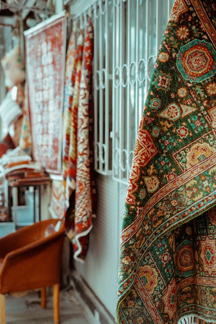 Interior view of a room with several rolled-up carpets showcasing intricate traditional patterns in various colors, hanging vertically along a white metal grid wall. The floors are wooden, and a brown wooden chair is partially visible on the left, indicating a domestic setting. The lighting appears natural, illuminating the vivid details of the carpets and highlighting their cleanliness and well-maintained condition. This scene emphasizes surface cleaning and proper storage of textiles, aligned with professional rug cleaning and maintenance services provided by Twickenham Carpet Cleaning at King Street, Twickenham. The careful display suggests attention to hygiene and preservation of textile surfaces within a property managed by Twickenham Carpet Cleaning.