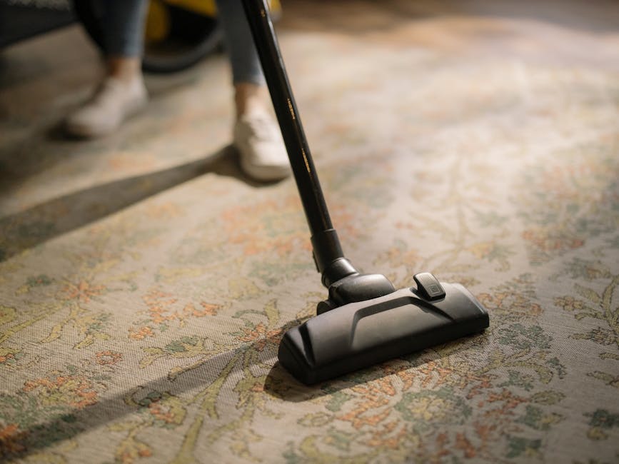A person vacuuming a detailed patterned carpet in a well-lit room, using a black and silver vacuum cleaner with a long handle. The carpet features intricate floral designs in muted tones of green, beige, pink, and orange. The individual is wearing casual clothing, including dark jeans and white sneakers, and is standing upright while cleaning. The clean and shiny surface of the carpet suggests professional deep cleaning by Twickenham Carpet Cleaning, aligning with best hygiene and maintenance practices in residential settings, especially relevant to Richmond Council's carpet cleaning rules highlighted on the webpage.