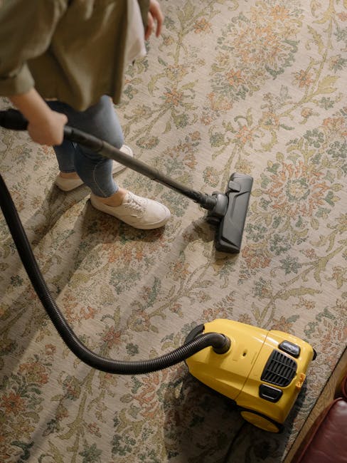 A person wearing beige shoes, blue jeans, and a beige coat uses a vacuum cleaner on a traditional patterned area rug with floral motifs in muted tones of beige, green, and orange. The vacuum cleaner is yellow with a black hose and a metal wand attachment, positioned in a well-lit room with wooden furniture nearby. Twickenham Carpet Cleaning specializes in surface cleaning and deep cleaning services, adhering to Richmond Council carpet cleaning rules, to ensure effective sanitisation and hygiene of residential carpets and rugs.